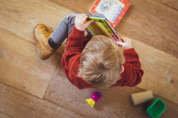 A toddler in a red sweater sitting on a wooden floor, absorbed in a picture book