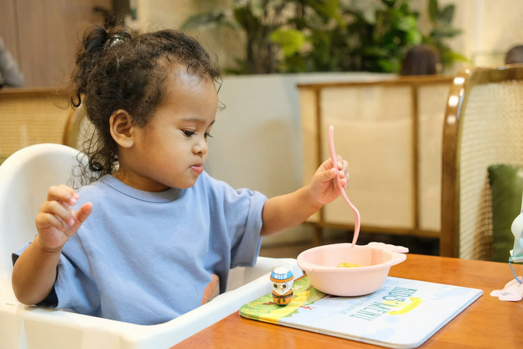 A toddler in a high chair feeding herself with a spoon and bowl